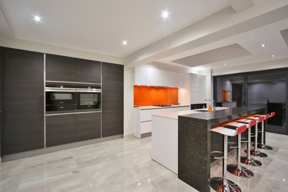 This is an example of a mid-sized contemporary single-wall open plan kitchen in London with an undermount sink, flat-panel cabinets, white cabinets, quartzite benchtops, orange splashback, glass sheet splashback, black appliances, porcelain floors, with island, beige floor and white benchtop.