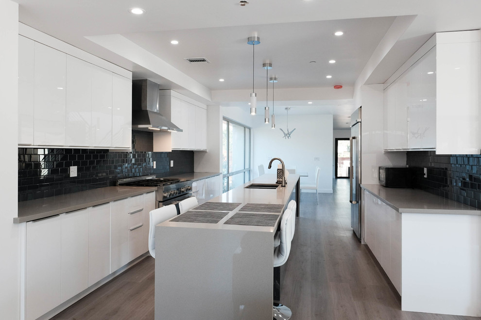 This is an example of a large modern galley eat-in kitchen in Los Angeles with an integrated sink, flat-panel cabinets, white cabinets, quartz benchtops, black splashback, ceramic splashback, stainless steel appliances, medium hardwood floors, with island and brown floor.