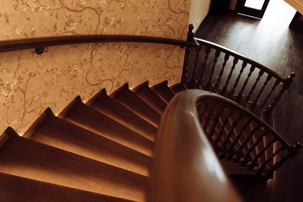 Стильный дизайн: большая изогнутая деревянная лестница в классическом стиле с деревянными ступенями, деревянными перилами, обоями на стенах и кладовкой или шкафом под ней - последний тренд