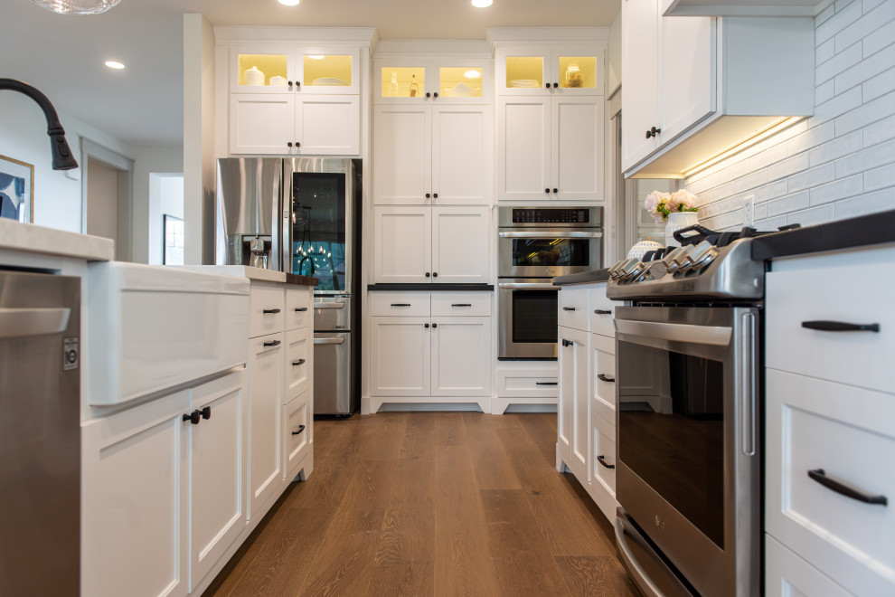 Large arts and crafts l-shaped open plan kitchen in Salt Lake City with a farmhouse sink, shaker cabinets, white cabinets, quartzite benchtops, white splashback, subway tile splashback, stainless steel appliances, medium hardwood floors, with island, brown floor and black benchtop.