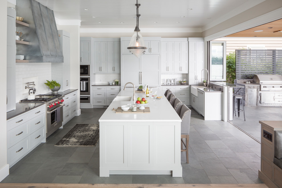 This is an example of a transitional u-shaped kitchen in Portland with a farmhouse sink, shaker cabinets, white cabinets, white splashback, subway tile splashback, panelled appliances, with island, grey floor and white benchtop.