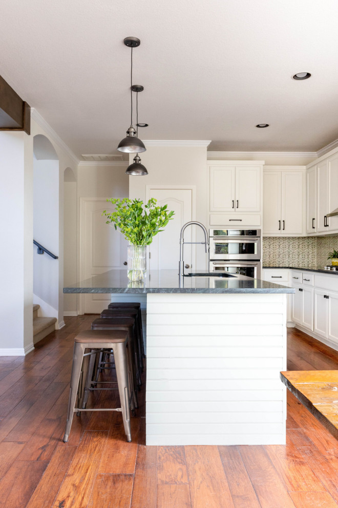 Eat-in kitchen - mid-sized transitional l-shaped medium tone wood floor and brown floor eat-in kitchen idea in Austin with an undermount sink, shaker cabinets, white cabinets, soapstone countertops, beige backsplash, ceramic backsplash, stainless steel appliances, an island and black countertops
