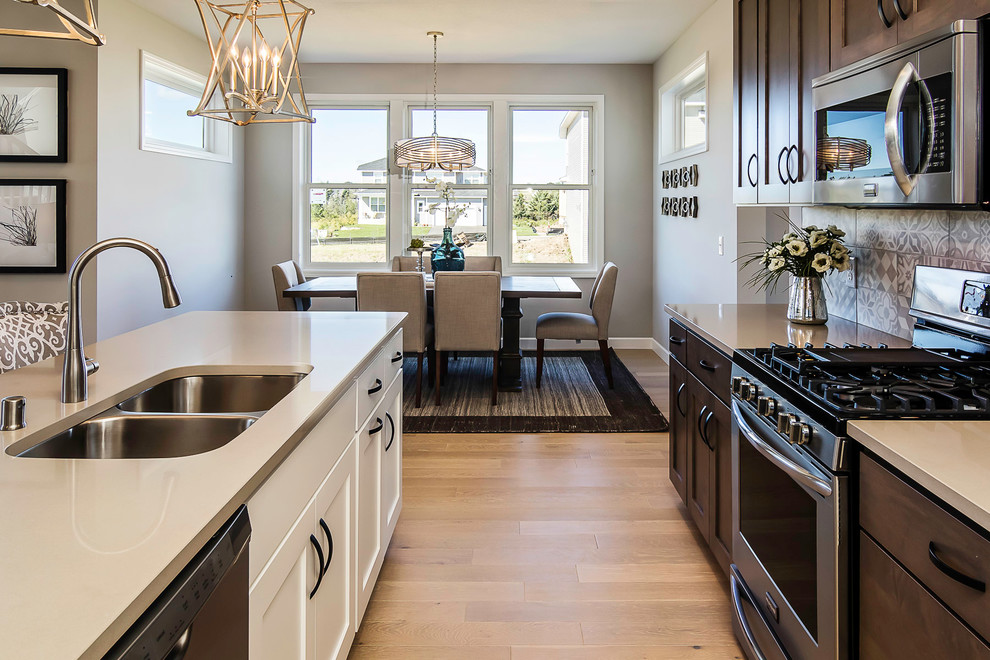Mid-sized transitional l-shaped eat-in kitchen in Minneapolis with a double-bowl sink, shaker cabinets, dark wood cabinets, solid surface benchtops, grey splashback, stainless steel appliances, light hardwood floors and with island.