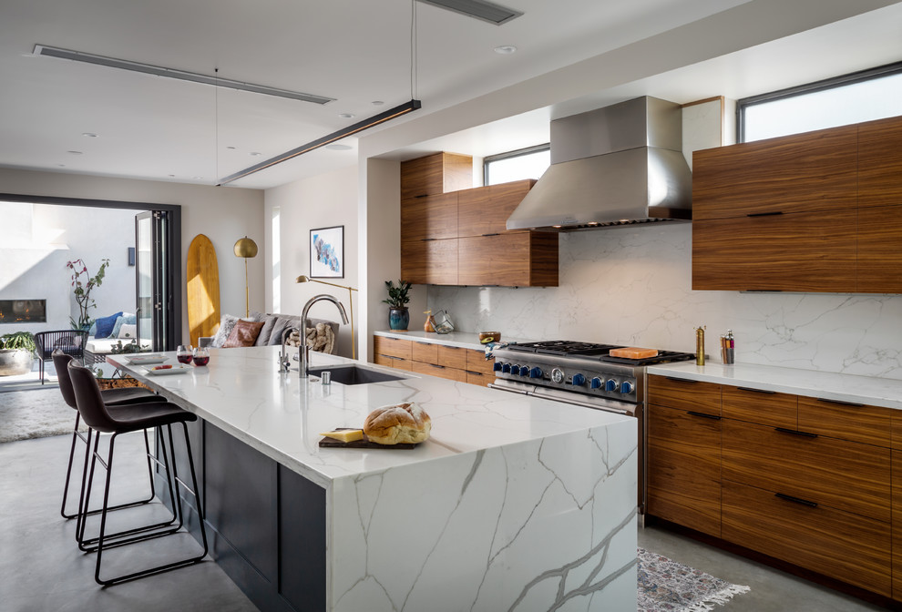 This is an example of a contemporary galley open plan kitchen in Orange County with an undermount sink, flat-panel cabinets, medium wood cabinets, white splashback, stone slab splashback, stainless steel appliances, concrete floors, with island, grey floor and white benchtop.