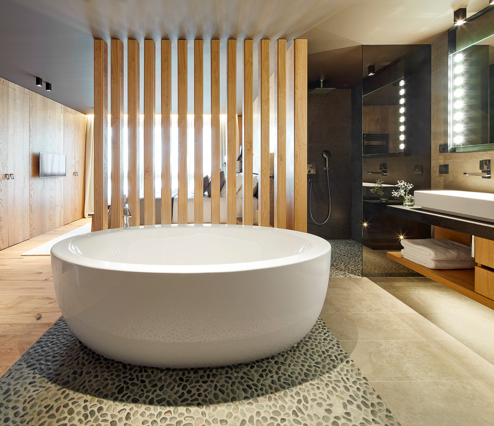 This is an example of a mid-sized modern master bathroom in Barcelona with flat-panel cabinets, medium wood cabinets, a freestanding tub, a curbless shower, a two-piece toilet, beige tile, pebble tile, grey walls, ceramic floors, a vessel sink, limestone benchtops, beige floor, an open shower and black benchtops.