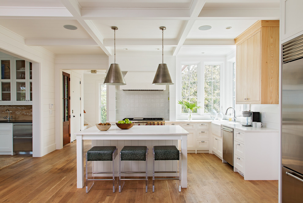 This is an example of a mid-sized beach style l-shaped kitchen in Charleston with a farmhouse sink, shaker cabinets, ceramic splashback, stainless steel appliances, with island, white cabinets, grey splashback and light hardwood floors.