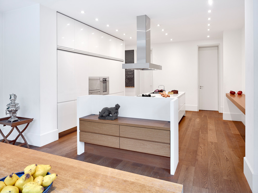 This is an example of an expansive contemporary eat-in kitchen in Frankfurt with a drop-in sink, flat-panel cabinets, white cabinets, marble benchtops, white splashback, stainless steel appliances, medium hardwood floors, with island, brown floor and white benchtop.