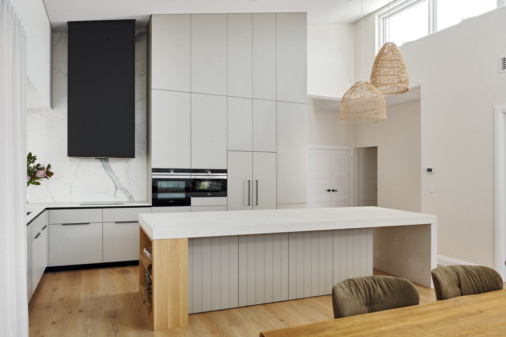 Design ideas for a large scandinavian l-shaped kitchen/diner in Perth with a double-bowl sink, flat-panel cabinets, grey cabinets, engineered stone countertops, white splashback, porcelain splashback, black appliances, light hardwood flooring, an island, beige floors, white worktops and a vaulted ceiling.