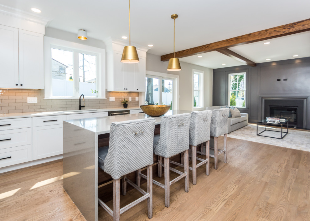 Example of a mid-sized transitional l-shaped light wood floor and white floor open concept kitchen design in Boston with an undermount sink, shaker cabinets, white cabinets, quartz countertops, gray backsplash, ceramic backsplash, stainless steel appliances, an island and white countertops