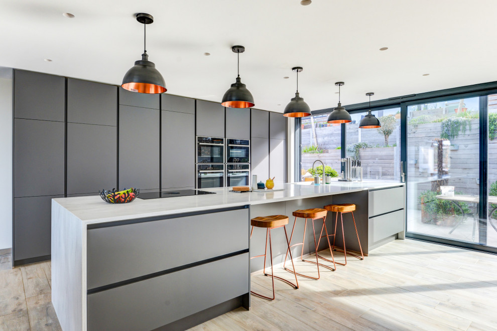 Design ideas for an expansive modern grey and white single-wall open plan kitchen in Sussex with an integrated sink, flat-panel cabinets, grey cabinets, composite countertops, black appliances, light hardwood flooring, an island, brown floors, grey worktops and feature lighting.