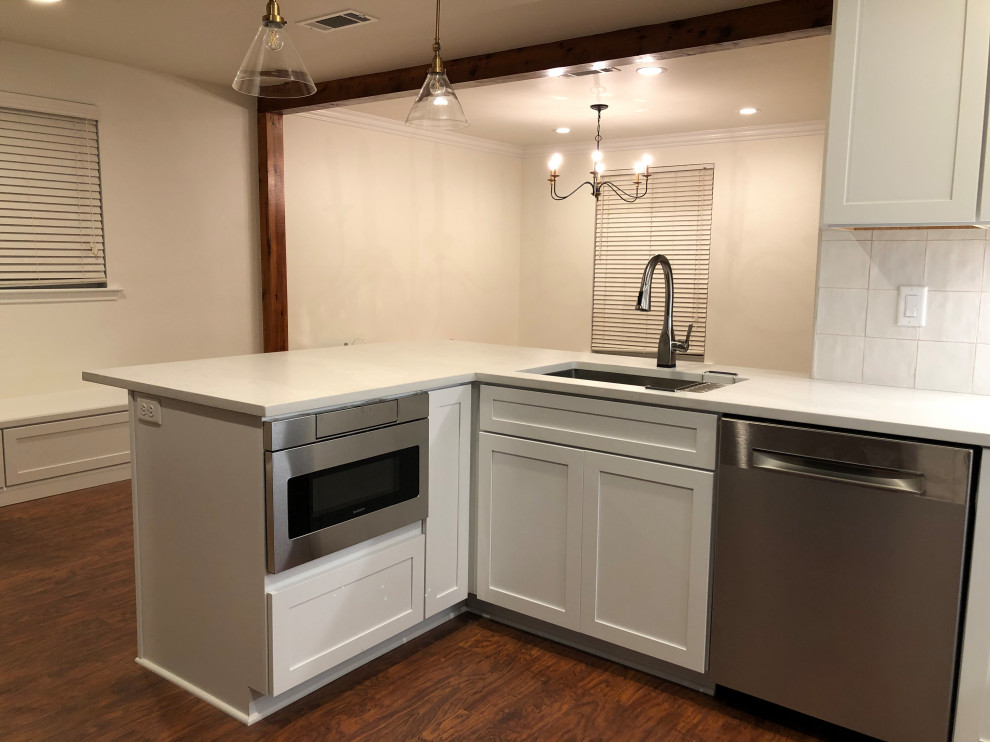 This is an example of a mid-sized transitional u-shaped open plan kitchen in New Orleans with an undermount sink, shaker cabinets, white cabinets, quartzite benchtops, white splashback, ceramic splashback, stainless steel appliances, medium hardwood floors, a peninsula, brown floor, white benchtop and exposed beam.