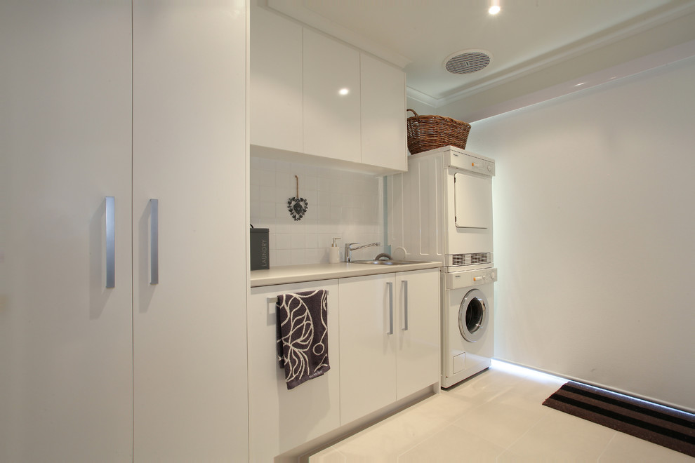 Large modern single-wall dedicated laundry room in Melbourne with a single-bowl sink, flat-panel cabinets, white cabinets, laminate benchtops, white walls, porcelain floors and a stacked washer and dryer.