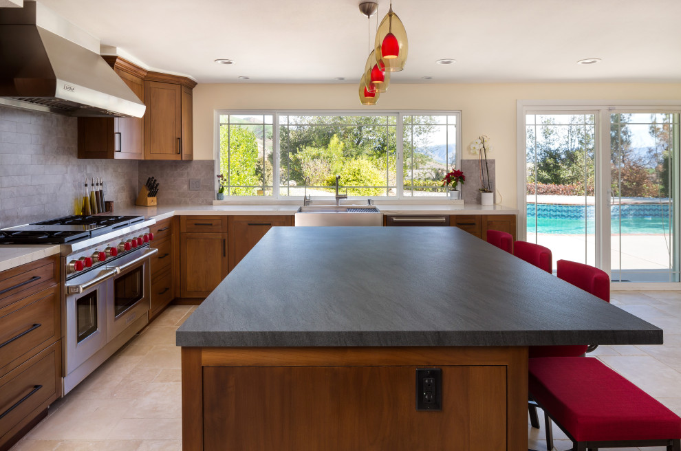 Photo of a mid-sized traditional l-shaped eat-in kitchen in Los Angeles with a farmhouse sink, raised-panel cabinets, medium wood cabinets, granite benchtops, grey splashback, porcelain splashback, stainless steel appliances, travertine floors, with island, multi-coloured floor and black benchtop.