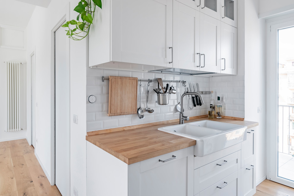 This is an example of a small scandinavian galley open plan kitchen in Rome with a double-bowl sink, flat-panel cabinets, white cabinets, wood benchtops, white splashback, ceramic splashback, stainless steel appliances, light hardwood floors and no island.