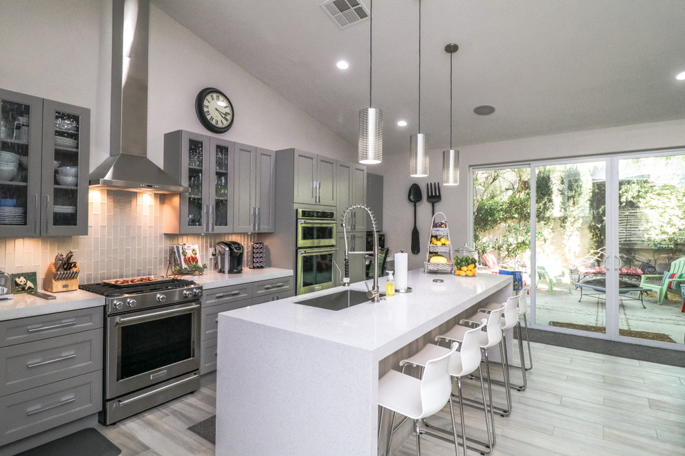 Photo of a large transitional l-shaped eat-in kitchen in Las Vegas with a single-bowl sink, recessed-panel cabinets, grey cabinets, quartzite benchtops, grey splashback, glass tile splashback, stainless steel appliances, light hardwood floors and with island.