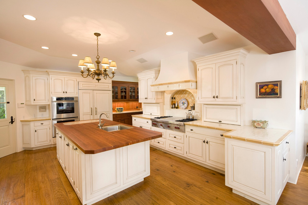 Design ideas for a large mediterranean l-shaped eat-in kitchen in Santa Barbara with an undermount sink, raised-panel cabinets, white cabinets, tile benchtops, panelled appliances, ceramic splashback, light hardwood floors, with island and beige splashback.