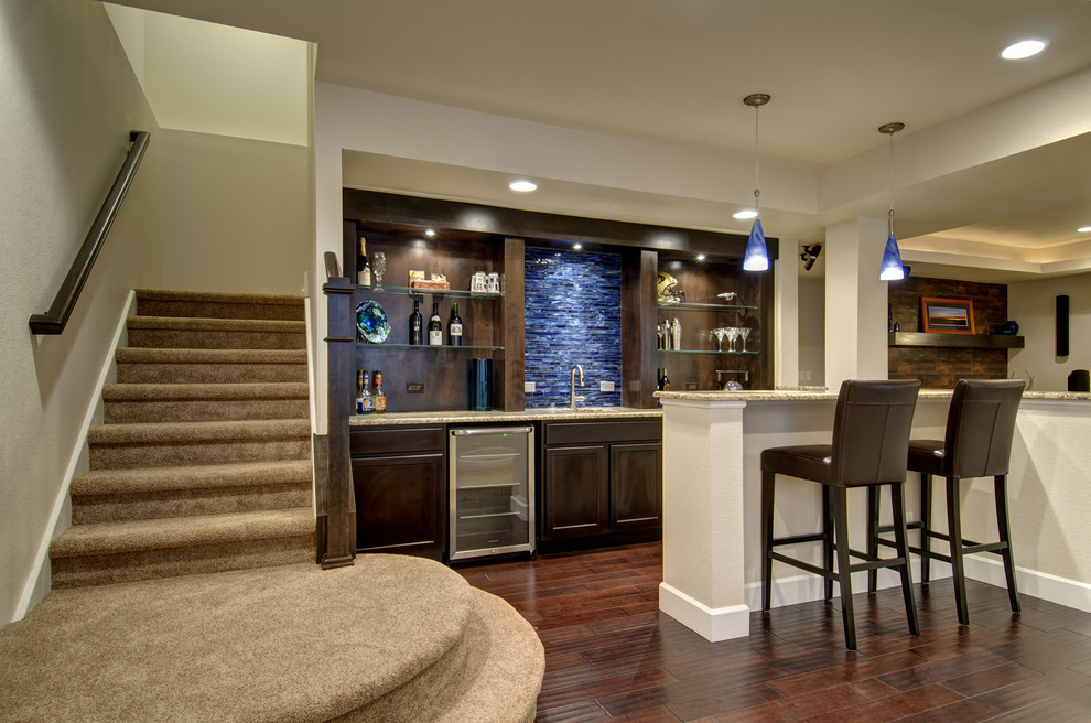 This is an example of a large transitional u-shaped seated home bar in Denver with an undermount sink, raised-panel cabinets, dark wood cabinets, granite benchtops, blue splashback, glass tile splashback, dark hardwood floors, brown floor and beige benchtop.