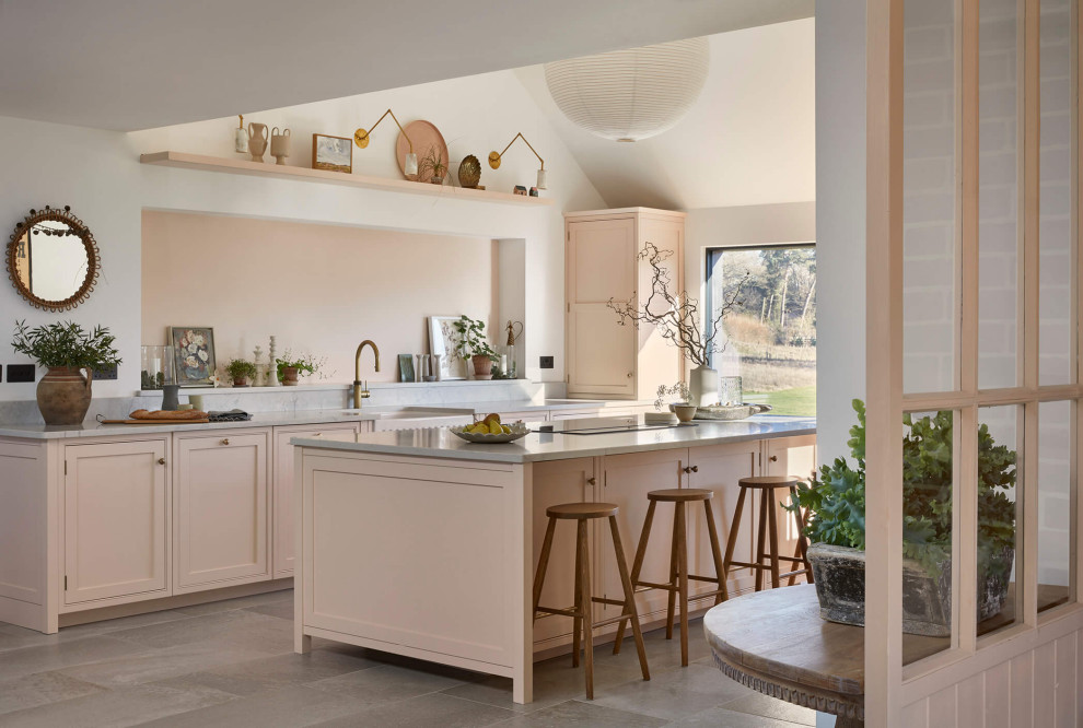 This is an example of a large country single-wall open plan kitchen in London with a farmhouse sink, shaker cabinets, pink cabinets, quartzite benchtops, pink splashback, black appliances, ceramic floors, with island, multi-coloured floor, grey benchtop and vaulted.