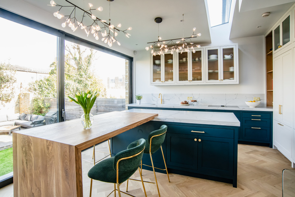 Inspiration for a large traditional l-shaped kitchen/diner in London with a built-in sink, shaker cabinets, blue cabinets, engineered stone countertops, white splashback, engineered quartz splashback, black appliances, light hardwood flooring, an island, brown floors and white worktops.