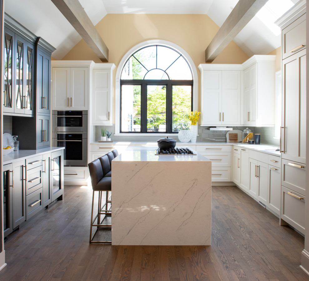 Large transitional u-shaped medium tone wood floor kitchen photo with an undermount sink, white cabinets, quartz countertops, gray backsplash, glass tile backsplash, stainless steel appliances, an island and white countertops