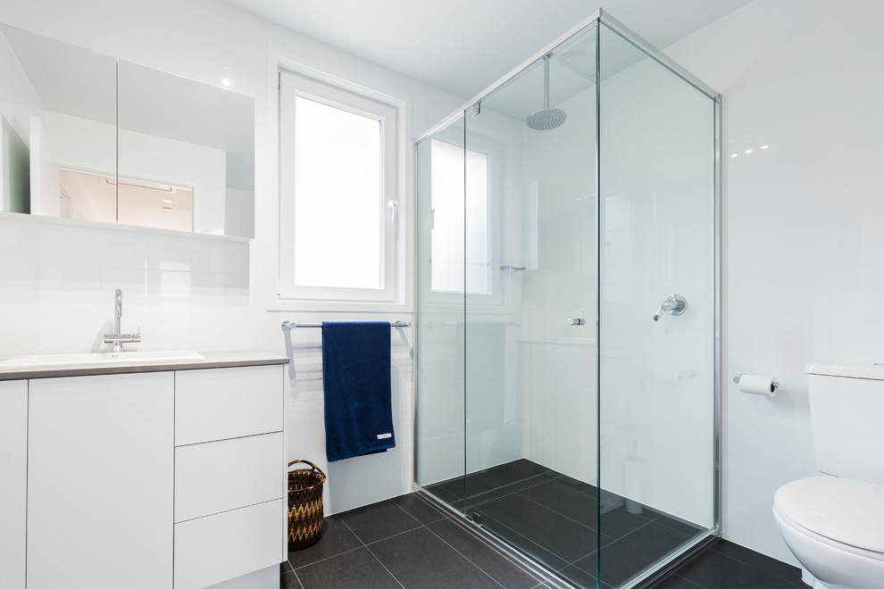 This is an example of a small contemporary master bathroom in Canberra - Queanbeyan with raised-panel cabinets, white cabinets, a corner shower, white tile, porcelain tile, white walls, porcelain floors, an integrated sink, laminate benchtops, grey floor, a hinged shower door and white benchtops.