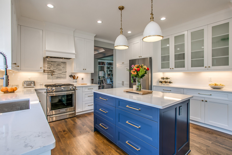 This is an example of a transitional u-shaped kitchen in Nashville with an undermount sink, shaker cabinets, white cabinets, white splashback, ceramic splashback, stainless steel appliances, dark hardwood floors, with island, brown floor and white benchtop.
