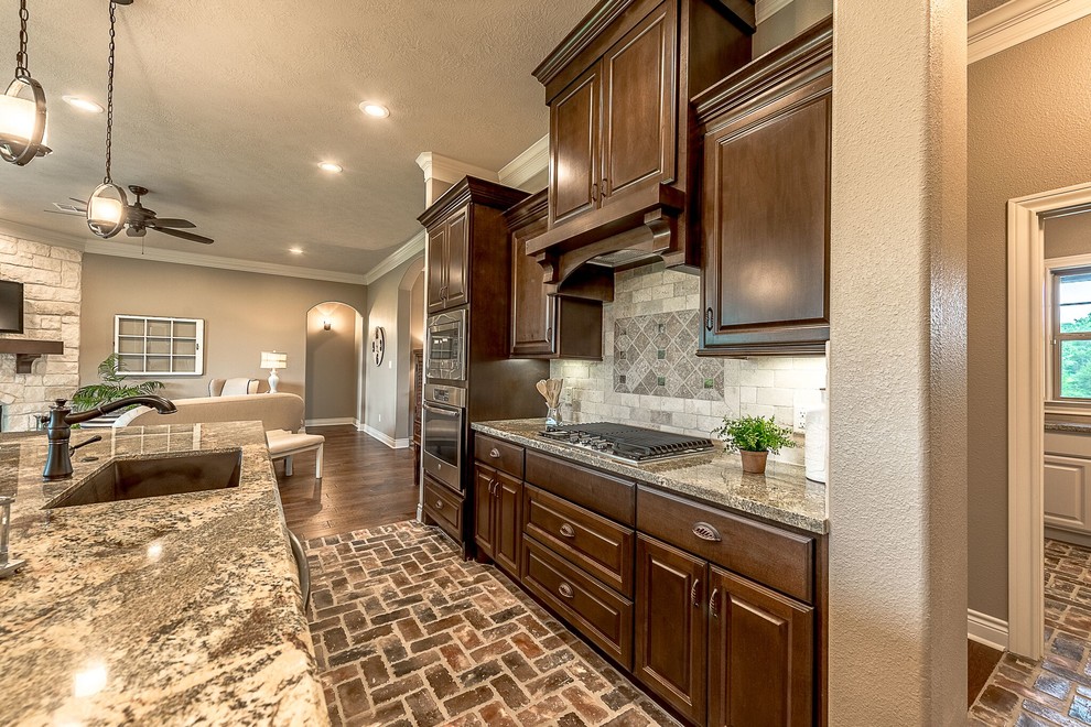 Open concept kitchen - mid-sized rustic u-shaped brick floor and brown floor open concept kitchen idea in Austin with a farmhouse sink, shaker cabinets, medium tone wood cabinets, granite countertops, beige backsplash, travertine backsplash, stainless steel appliances and an island