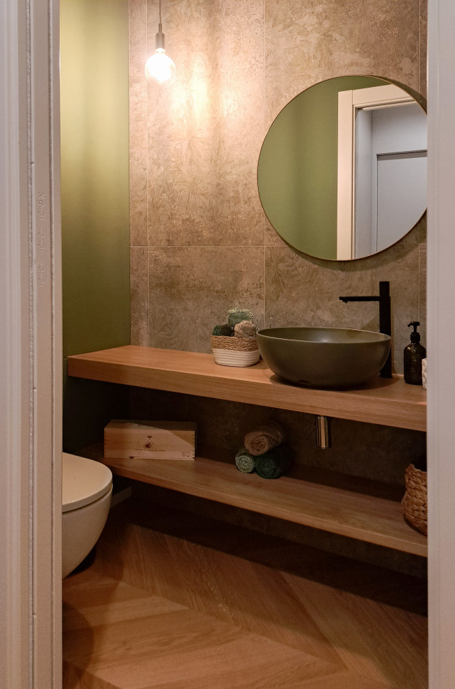 This is an example of a mid-sized contemporary powder room in Other with light wood cabinets, a wall-mount toilet, green tile, porcelain tile, green walls, light hardwood floors, a vessel sink, wood benchtops and a floating vanity.