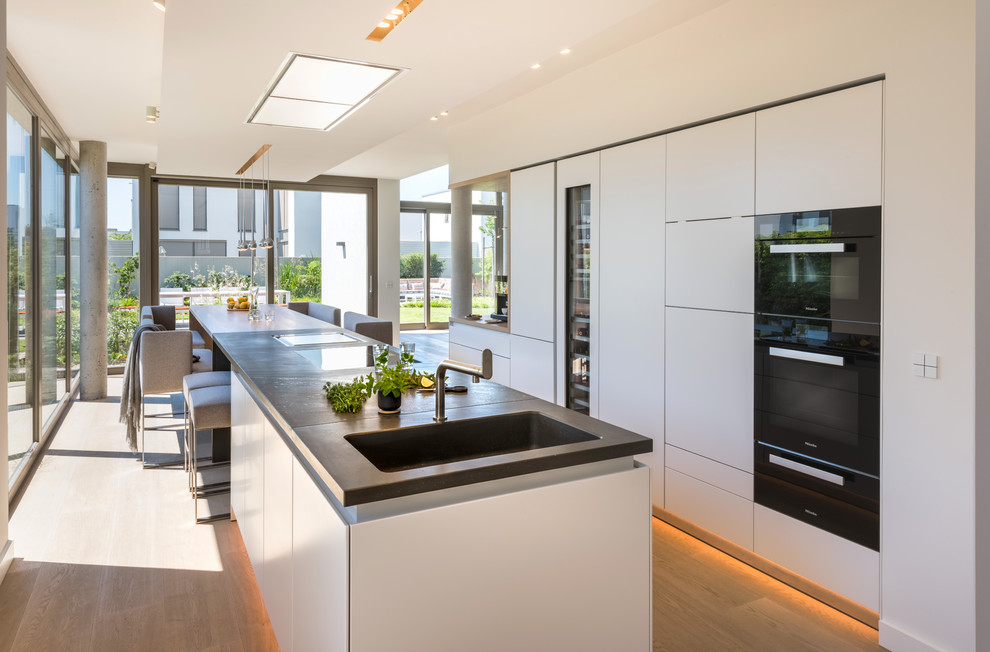 This is an example of a large contemporary galley eat-in kitchen in Cologne with an integrated sink, flat-panel cabinets, white cabinets, concrete benchtops, black appliances, medium hardwood floors, multiple islands and brown floor.