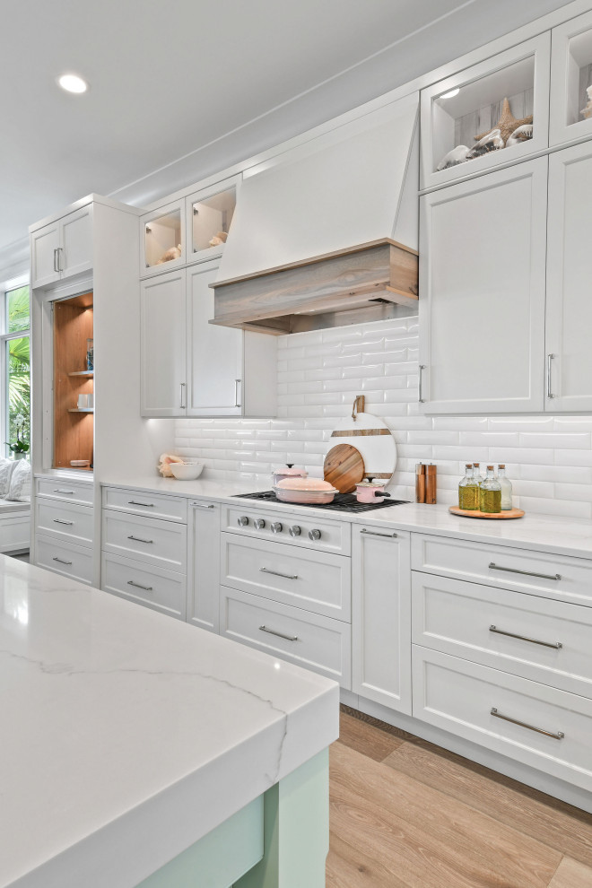 Photo of a mid-sized beach style l-shaped kitchen in Other with white cabinets, quartz benchtops, white splashback, subway tile splashback, panelled appliances, light hardwood floors, with island, brown floor, white benchtop and recessed.