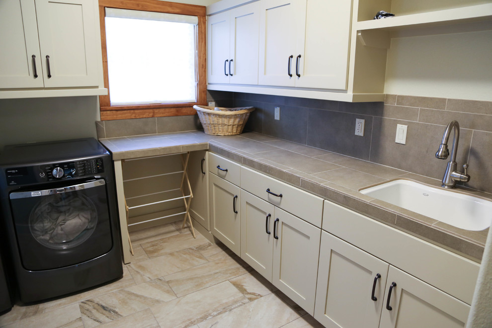 This is an example of a mid-sized contemporary l-shaped dedicated laundry room in Austin with a side-by-side washer and dryer, a drop-in sink, shaker cabinets, beige cabinets, tile benchtops, beige walls and travertine floors.