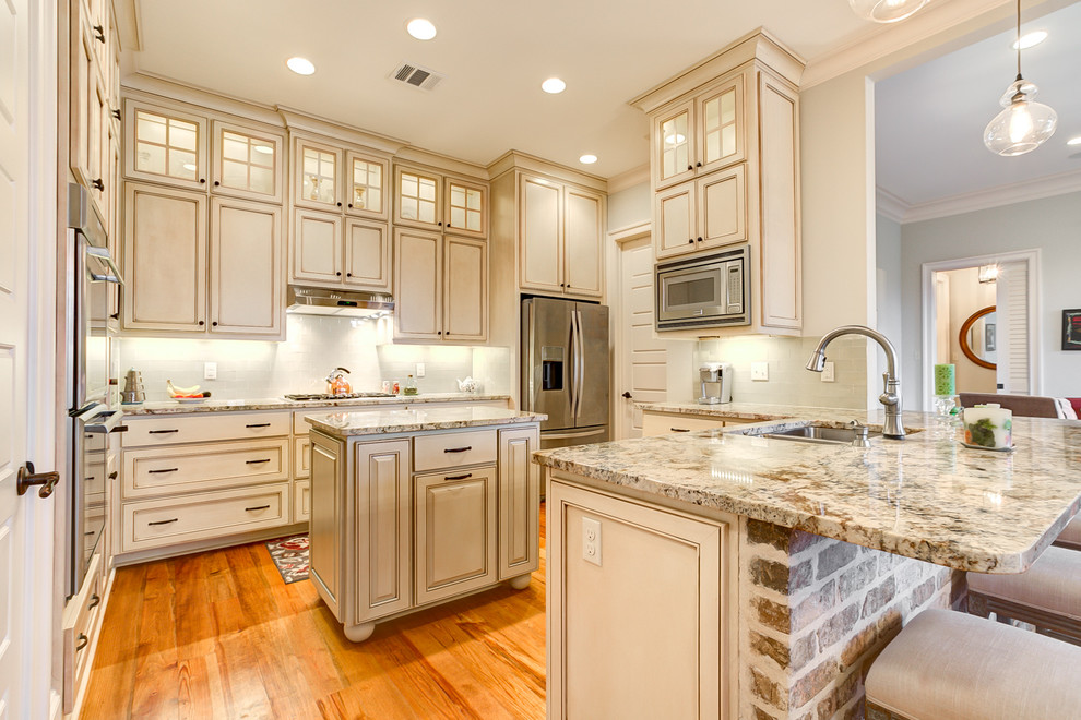 Photo of a mid-sized country u-shaped open plan kitchen in New Orleans with an undermount sink, beaded inset cabinets, beige cabinets, granite benchtops, glass tile splashback, stainless steel appliances, medium hardwood floors and with island.