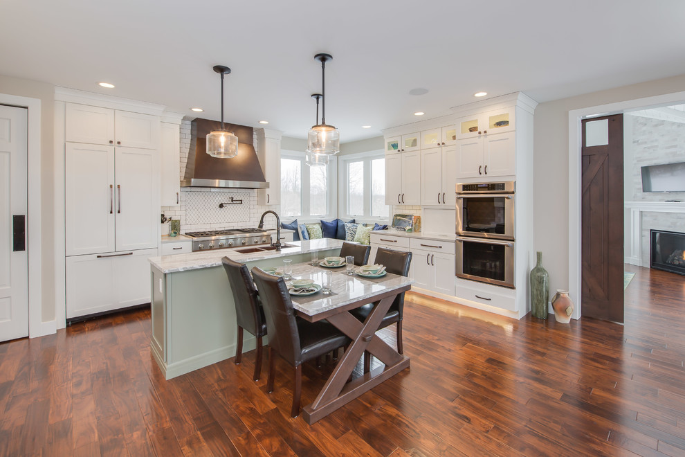 Photo of a large transitional l-shaped eat-in kitchen in Grand Rapids with white cabinets, quartz benchtops, white splashback, ceramic splashback, stainless steel appliances, medium hardwood floors, with island, brown floor, a farmhouse sink and shaker cabinets.