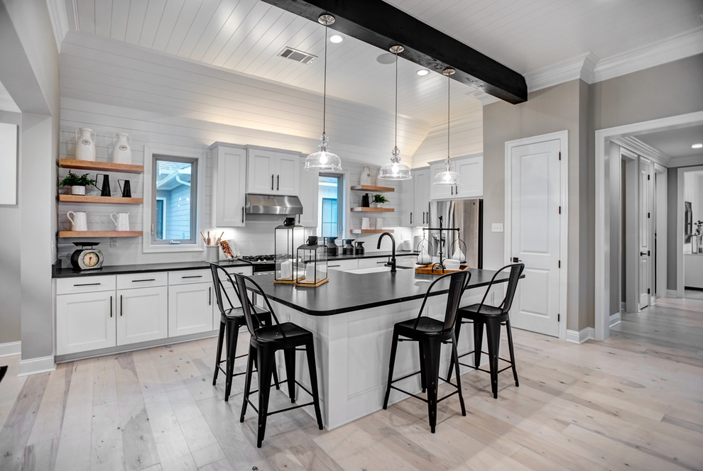 Country l-shaped kitchen with a farmhouse sink, shaker cabinets, white cabinets, stainless steel appliances, light hardwood floors, with island, beige floor and black benchtop.