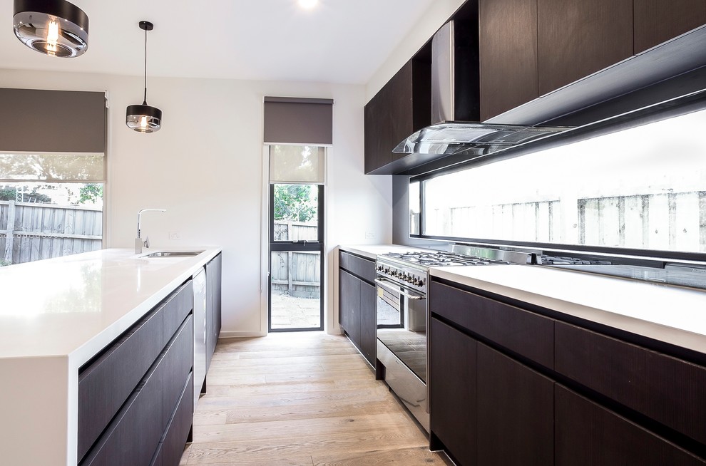 Mid-sized contemporary galley open plan kitchen in Melbourne with an undermount sink, dark wood cabinets, quartz benchtops, glass sheet splashback, stainless steel appliances, light hardwood floors, with island, brown floor and white benchtop.