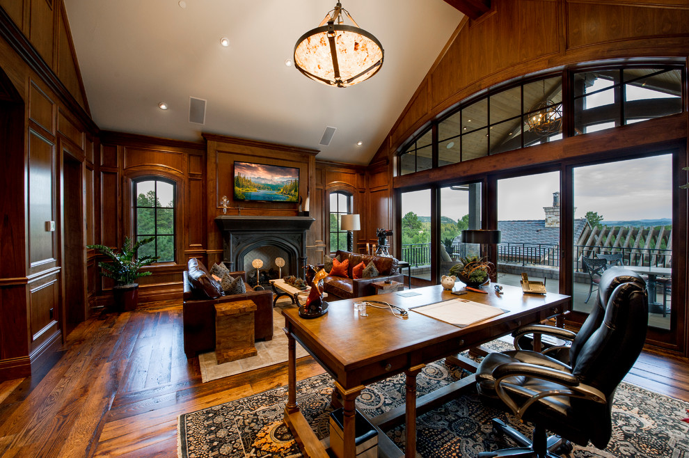 Photo of a large country study room in Denver with medium hardwood floors, a freestanding desk, brown walls, a standard fireplace, a wood fireplace surround and brown floor.