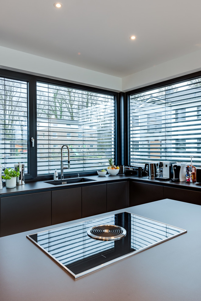 This is an example of a mid-sized contemporary open plan kitchen in Hamburg with a drop-in sink, flat-panel cabinets, grey cabinets, quartz benchtops, grey splashback, window splashback, black appliances, light hardwood floors, with island, beige floor and grey benchtop.