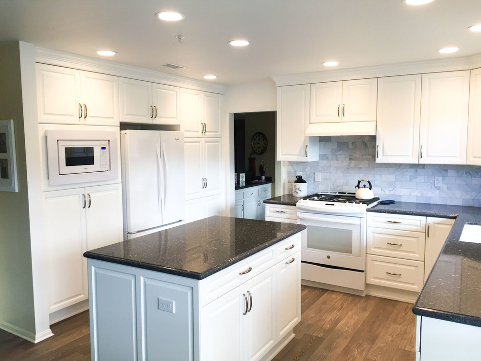 This is an example of a mid-sized traditional u-shaped kitchen in Los Angeles with an undermount sink, raised-panel cabinets, white cabinets, granite benchtops, grey splashback, marble splashback, white appliances, medium hardwood floors, with island, brown floor and black benchtop.