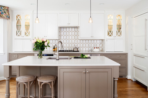 Two-tone kitchen with white glass-door uppers and taupe Shaker RTA cabinets, quartz island with stools, and arabesque tile backsplash.
