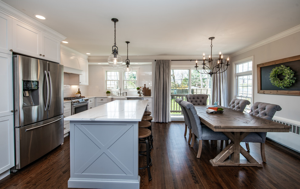 This is an example of a mid-sized country l-shaped eat-in kitchen in New York with a farmhouse sink, shaker cabinets, white cabinets, quartz benchtops, white splashback, subway tile splashback, stainless steel appliances, dark hardwood floors, with island, brown floor and white benchtop.