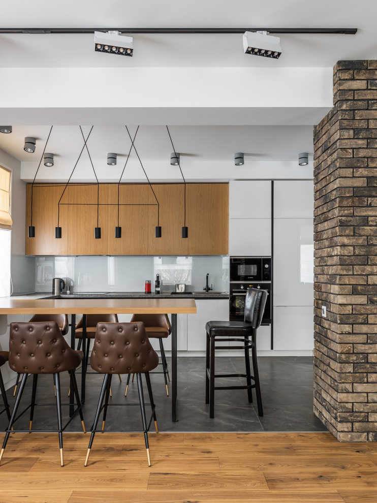Inspiration for a mid-sized contemporary l-shaped eat-in kitchen in Other with a drop-in sink, flat-panel cabinets, white cabinets, white splashback, glass sheet splashback, panelled appliances, grey floor and black benchtop.