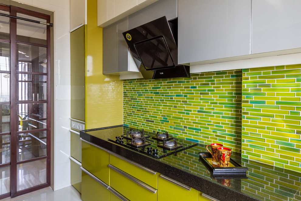 This is an example of a contemporary kitchen in Mumbai with flat-panel cabinets, green cabinets, green splashback, glass tile splashback, black appliances, white floor and black benchtop.