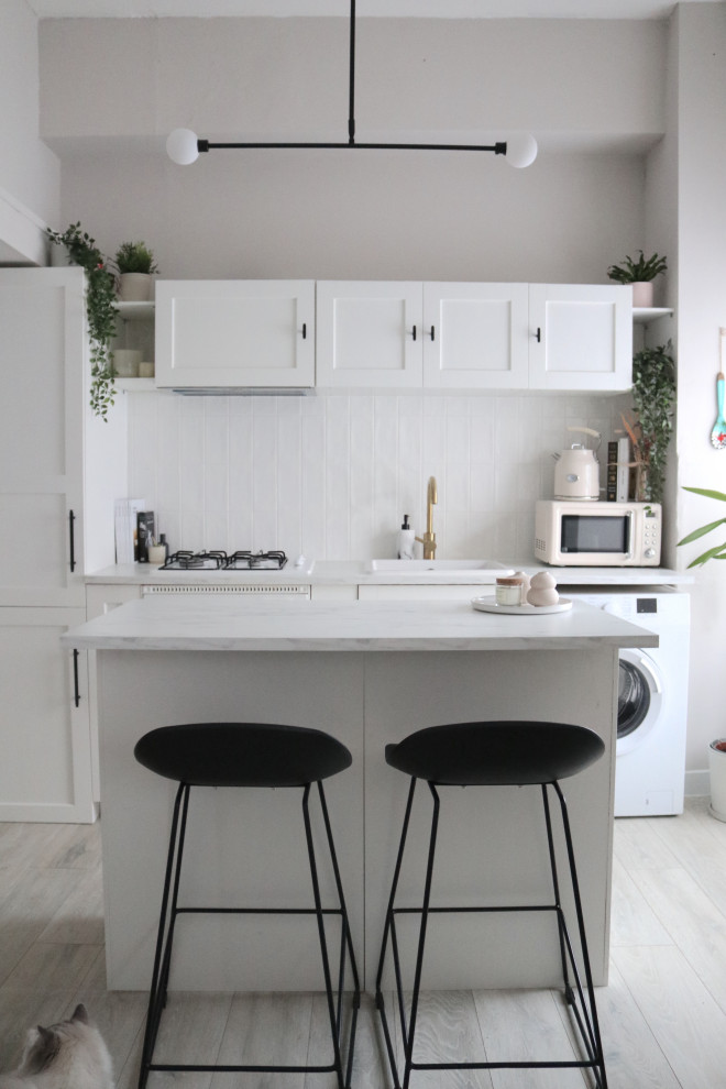 Foto på ett litet funkis vit linjärt kök med öppen planlösning, med en undermonterad diskho, skåp i shakerstil, vita skåp, laminatbänkskiva, vitt stänkskydd, stänkskydd i keramik, vita vitvaror, vinylgolv, en köksö och beiget golv