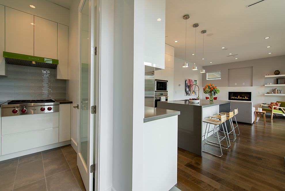 This is an example of a mid-sized modern l-shaped open plan kitchen in Vancouver with an undermount sink, flat-panel cabinets, white cabinets, solid surface benchtops, grey splashback, stainless steel appliances, medium hardwood floors, with island, brown floor and grey benchtop.