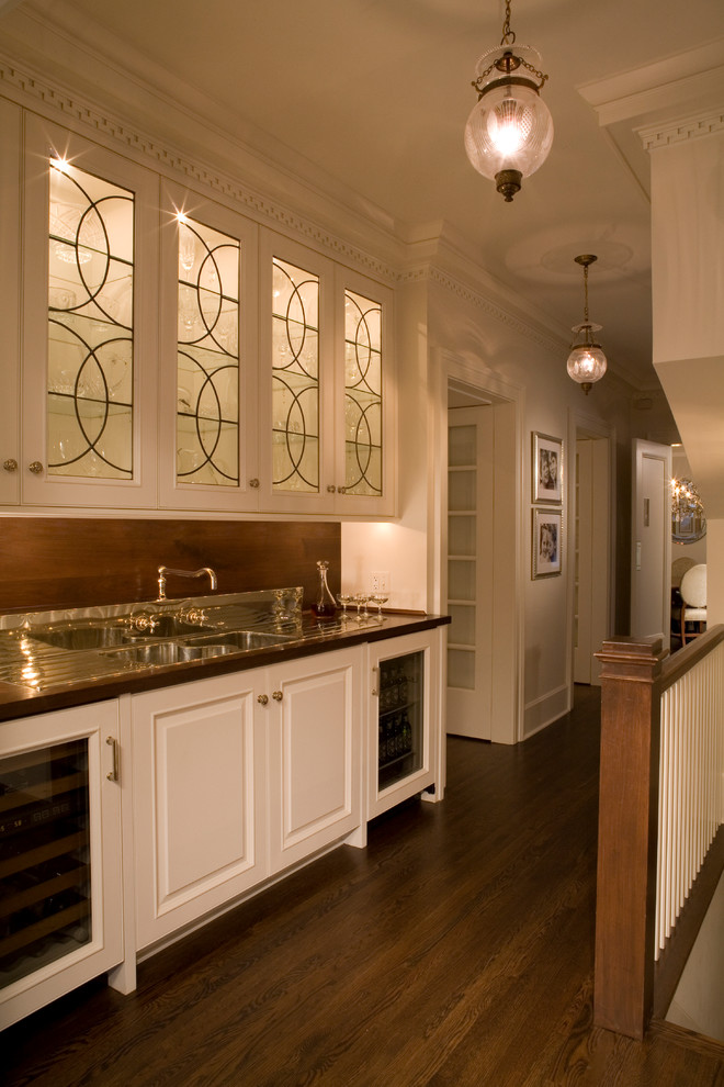 This is an example of a mid-sized traditional single-wall wet bar in Chicago with an undermount sink, glass-front cabinets, white cabinets, wood benchtops, brown splashback, timber splashback and dark hardwood floors.