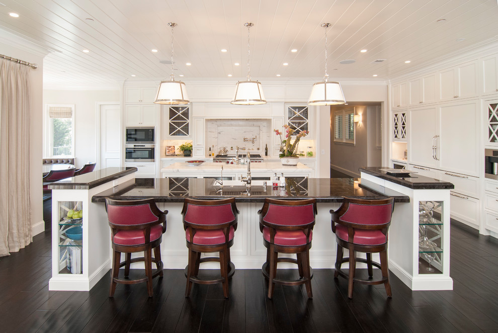 This is an example of a traditional eat-in kitchen in San Francisco with a farmhouse sink, shaker cabinets, white cabinets, white splashback, panelled appliances, dark hardwood floors, multiple islands and brown floor.