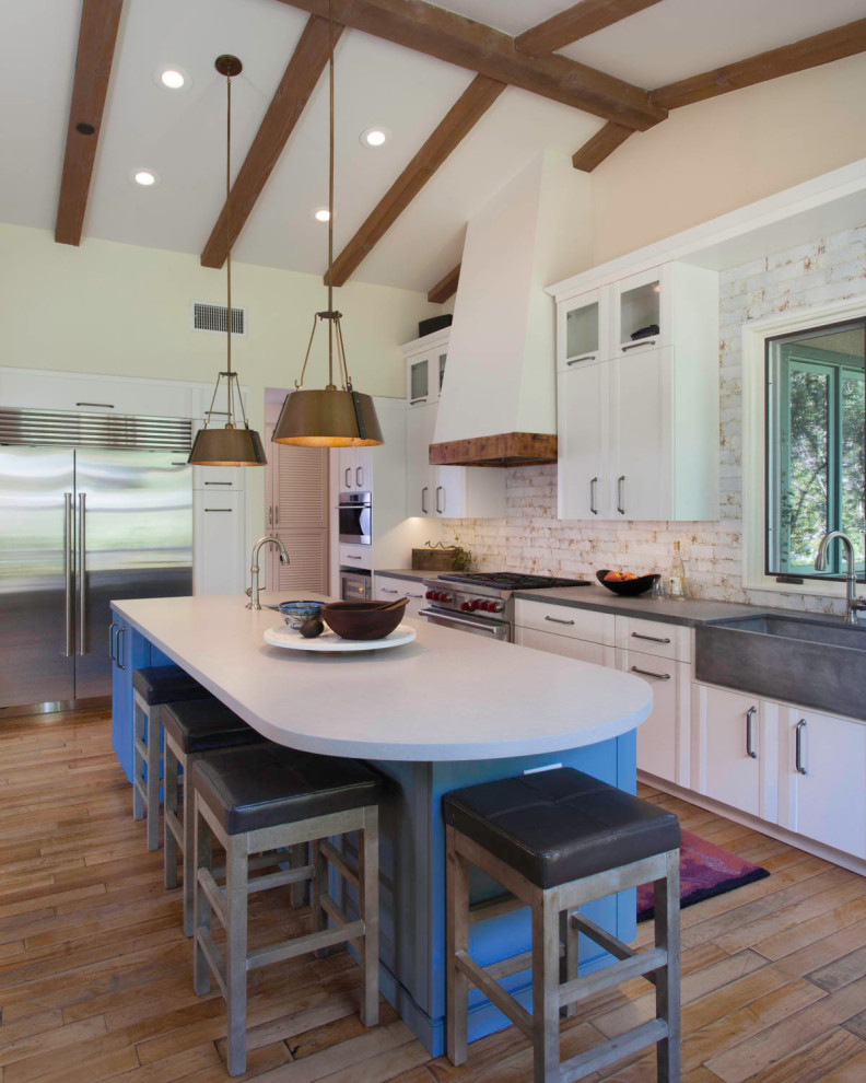 Photo of a large country l-shaped open plan kitchen in Denver with a farmhouse sink, recessed-panel cabinets, white cabinets, quartz benchtops, white splashback, ceramic splashback, stainless steel appliances, light hardwood floors, with island, brown floor, grey benchtop and exposed beam.