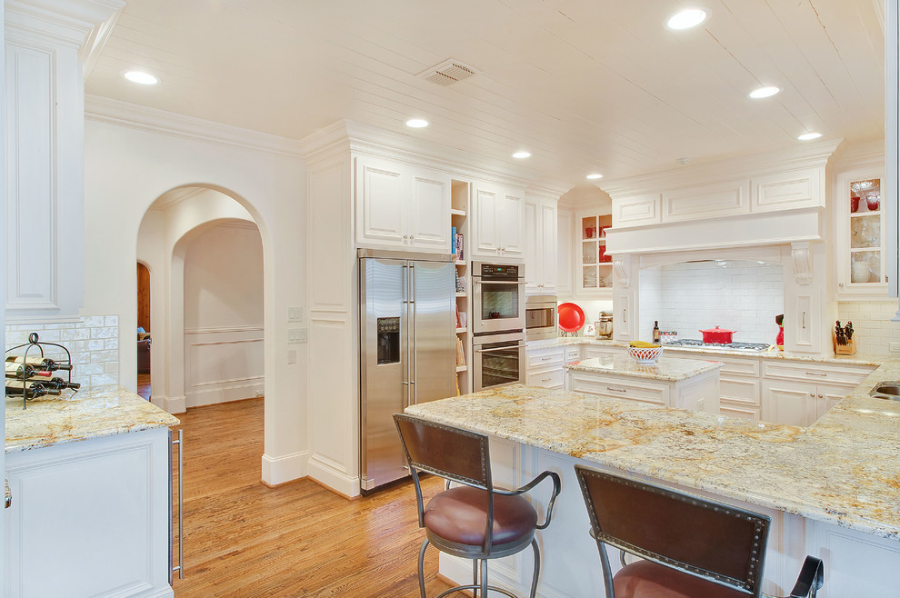 This is an example of a traditional u-shaped kitchen in Dallas with a double-bowl sink, raised-panel cabinets, white cabinets, granite benchtops, white splashback, subway tile splashback, stainless steel appliances, medium hardwood floors and with island.