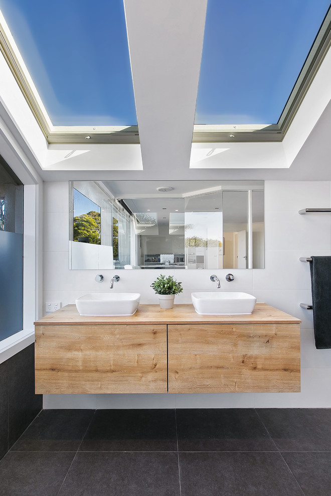 Design ideas for a contemporary master bathroom in Melbourne with a freestanding tub, an open shower, a wall-mount toilet, gray tile, porcelain tile, white walls, porcelain floors, a vessel sink, wood benchtops, flat-panel cabinets, light wood cabinets and brown benchtops.