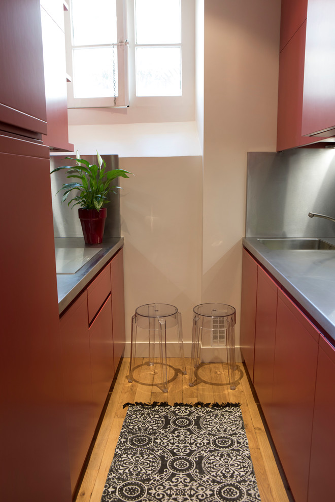 Small contemporary galley separate kitchen in Paris with an integrated sink, beaded inset cabinets, red cabinets, stainless steel benchtops, grey splashback, panelled appliances, light hardwood floors, brown floor and grey benchtop.